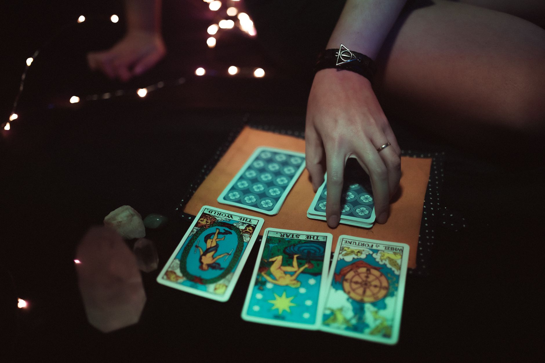 Unrecognizable female reading tarot cards while sitting on floor in dark room with glowing bright lights and crystals at home